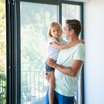 Cheerful father holding cute daughter, looking at her and smiling. Lovely blonde girl looking at camera. Happy dad with kid standing near balcony open door. Relocation and family moving day concept